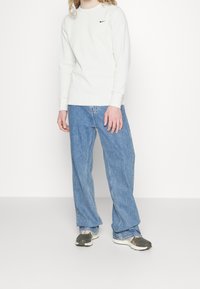 White long-sleeve ribbed shirt with a small black Nike logo, paired with light blue wide-leg jeans and grey sneakers with white soles.