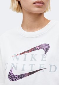 Κοντινό πλάνο ενός ατόμου που φοράει λευκό μπλουζάκι Nike με διπλά λογότυπα Swoosh με λεοπάρ σχέδιο σε μοβ και κόκκινο και το κείμενο "NIKE UNITED".