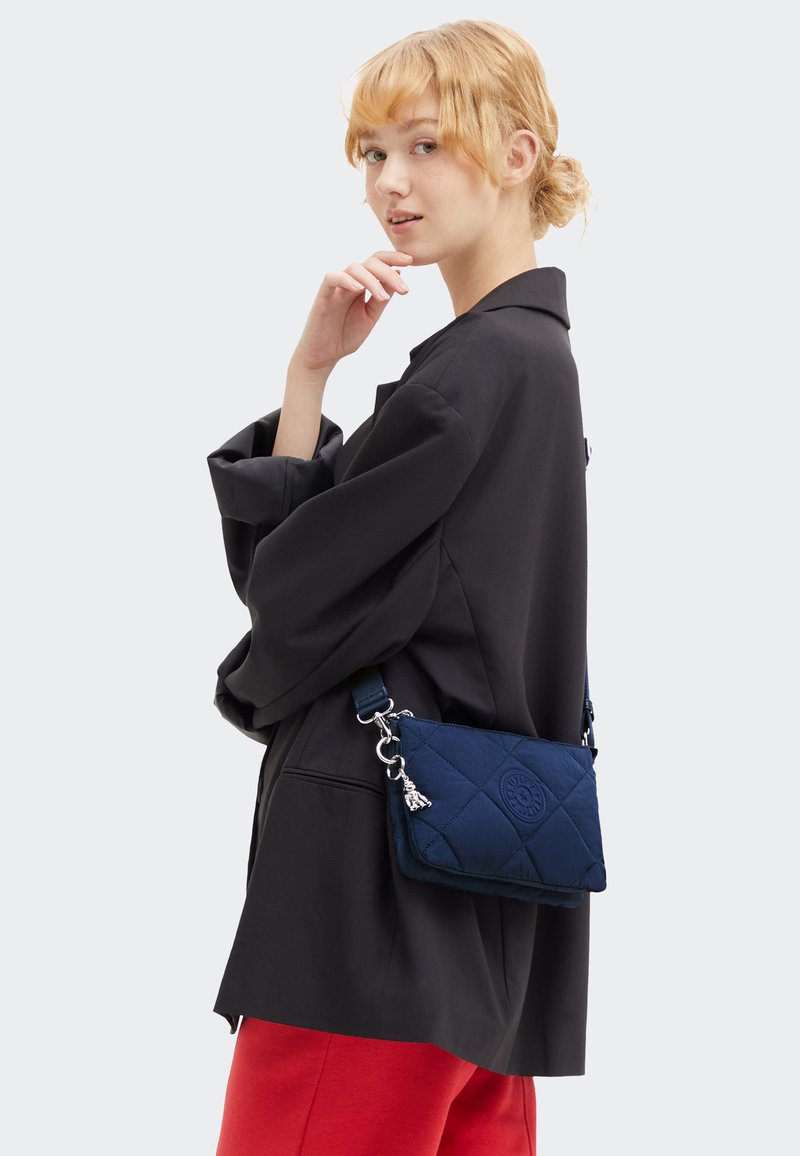 Borsa a tracolla trapuntata blu navy con tracolla removibile, caratterizzata da un dettaglio con logo e accessori in argento. Indossata di lato da una persona in giacca nera.