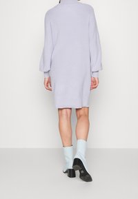 Abito in maglia oversized di colore lilla con collo alto, maniche lunghe a sbuffo e texture a coste. Abbinato a stivaletti azzurri e tacchi neri.