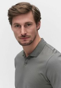 Grå polo med strukturerad väv, med krage och korta ärmar. Modellen har kort hår och välansad skägg.