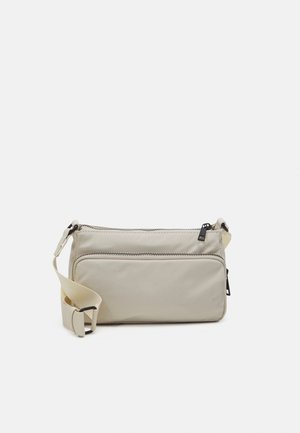 Beige crossbodytas met verstelbare riem, twee met rits afgesloten vakken en zwarte ritstrekkers, afgebeeld op een effen witte achtergrond.