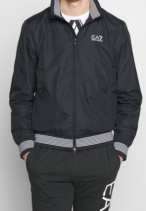 Man draagt een zwarte EA7 Emporio Armani jas met rits en bijpassende zwarte broek met witte gestreepte boorden en logodetails.