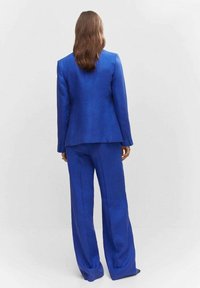 Costume bleu sur mesure avec un blazer structuré et un pantalon à jambes larges, fabriqué en tissu lisse. Présente des détails subtils sur les manches et le dos.