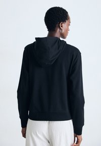 Persoon met kort krullend haar, gekleed in een zwart hoodie en witte, gestructureerde broek, met de rug naar de camera gekeerd tegen een eenvoudige lichte achtergrond.