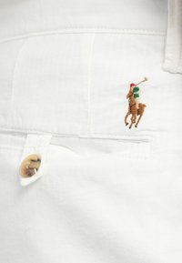 Materialul din bumbac alb prezintă un mic design brodat cu un jucător de polo în culorile roșu și verde pe un cal, având un detaliu cu un nasture bej.