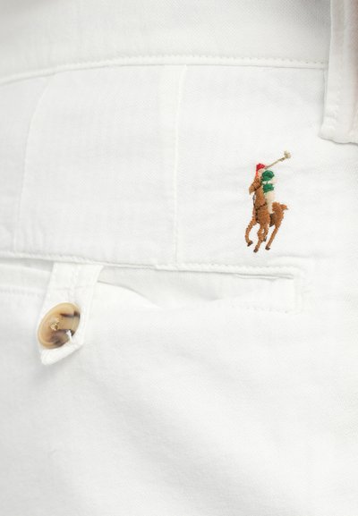 Hvit bomullsstoff har et lite brodert motiv av en polo-spiller i rødt og grønt på en hest, med en beige knappdetalj.