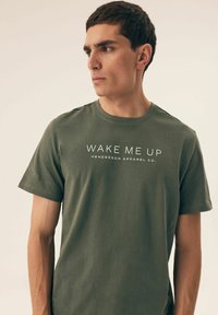 Oliwkowa bawełniana koszulka z okrągłym dekoltem, krótkimi rękawami i białym napisem "WAKE ME UP HENDERSON APPAREL CO." na przodzie.