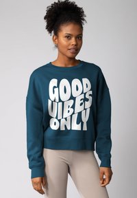 Teal sweatshirt met de tekst "GOOD VIBES ONLY" in grote witte letters. Heeft een ronde hals en verlaagde schouders. Gecombineerd met lichte geribde legging.