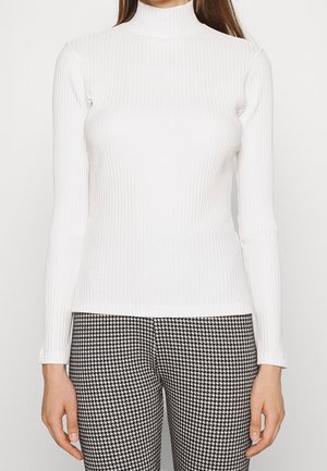 Frau trägt einen weißen, gerippten Rollkragenpullover mit langen Ärmeln und schwarz-weiß karierten Hosen im Hahnentrittmuster, steht vor einem schlichten Hintergrund.