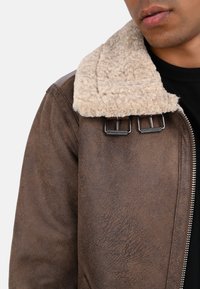 Hnědá bunda z umělé kůže s texturovaným povrchem, zdobená béžovým límcem z ovčí vlny a kovovými akcenty. Zapínání na zip vpředu.