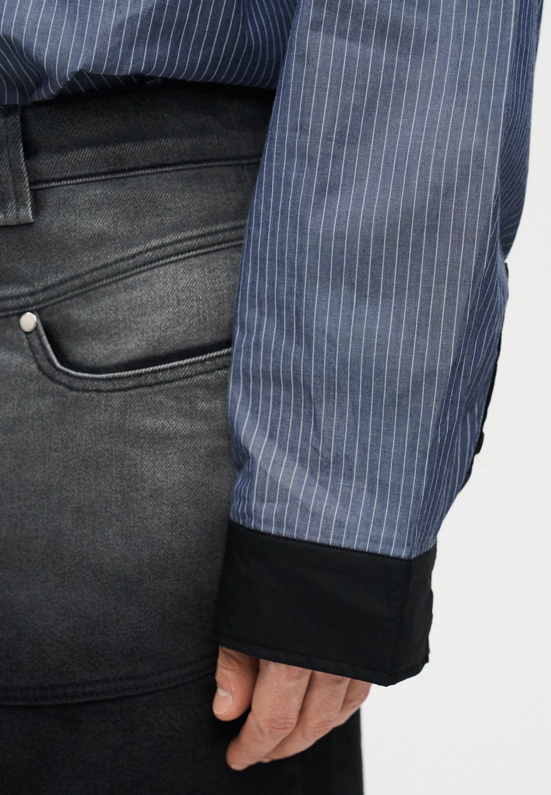 Sleeve of a blue pinstriped shirt with black cuff resting beside black faded denim jeans pocket.