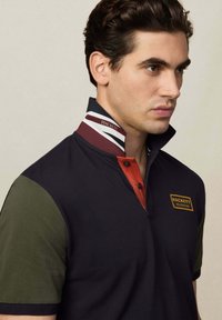 Námornícky polo tričko s červeným golierom a zelenými rukávmi, s vyšitým logom. Golier má pruhy a vyvýšený dizajnový detail.