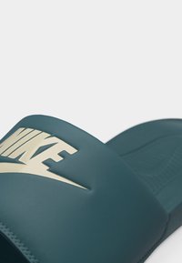 Artimo žalios spalvos Nike šlepetės, kurių dirželyje yra rusvos spalvos Nike logotipas.