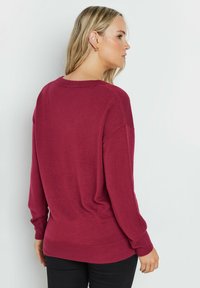 Bordeauxfarbener Oversized-Pullover mit langen Ärmeln, gerippten Bündchen und Saum. Weiche Textur sichtbar, mit weitem Ausschnitt und lockerer Passform.