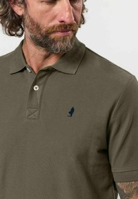 Homme aux cheveux bouclés et à la barbe portant un polo vert olive avec un petit logo bleu foncé sur la poitrine, regardant vers le bas et sur le côté.