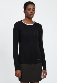 Jersey negro de manga larga con un diseño de punto trenzado texturizado, cuello redondo y dobladillo acanalado. El modelo lleva una camisa blanca debajo.