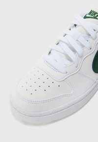 Baskets blanches avec un bout perforé, accent de logo vert, lacets plats blancs et semelle en caoutchouc texturée. Tige en cuir lisse avec des coutures contrastées.