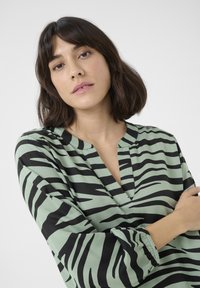 Žalia bliuska su juodais zebra dryžiais, V formos iškirpte, ilgomis rankovėmis su elastiniais riešais ir sklandžiu, lengvu audinio tekstūru.