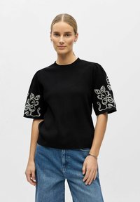 Femme aux cheveux blonds attachés en queue de cheval, portant un pull noir à manches courtes avec une broderie florale blanche, un jean bleu et des boucles d'oreilles créoles en argent.