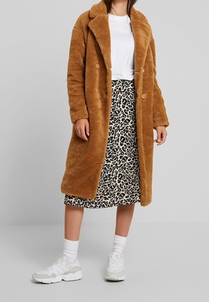 Manteau en fausse fourrure marron, croisé avec de grands boutons, associé à une jupe imprimé animal noir et blanc longueur genoux et des baskets blanches.