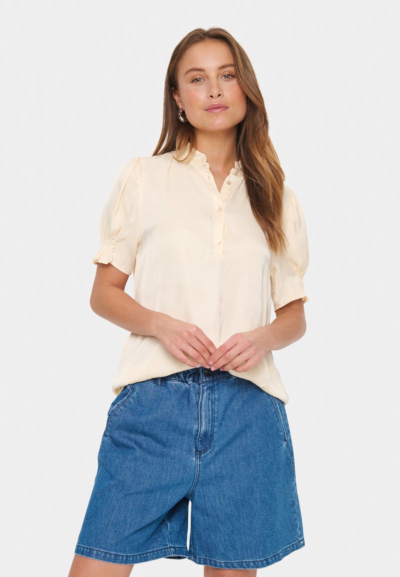 Lichtbeige blouse met knoopsluiting, ruches-kraag en opgezette mouwen, gecombineerd met donkerblauwe denim shorts.