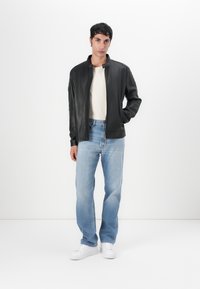 Chaqueta de cuero negra con cierre de cremallera, llevada sobre una camisa de color beige claro, combinada con jeans de mezclilla azul claro y zapatillas blancas.