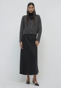 Grauer, kurz geschnittener Cardigan mit Knöpfen, kombiniert mit einem langen schwarzen Satinrock und schwarzen spitzen Absätzen. Glatte Texturen und schlichte Linien.