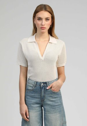 Femme vêtue d'un haut en maille blanc à manches courtes avec col et d'un jean taille haute bleu clair, debout avec une main dans la poche devant un fond uni.
