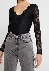 Body in pizzo nero con maniche lunghe, scollatura a V profonda, motivo floreale; abbinato a jeans in denim grigio chiaro a vita alta.