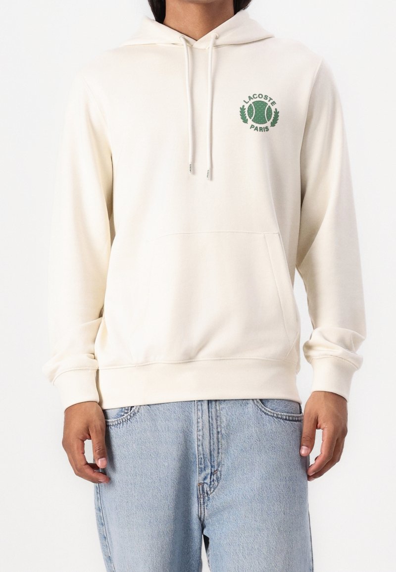 Krémová mikina s kapucí, klokaní kapsou, šňůrkou na kapuci a zeleným logem na hrudi. Měkká tkanina s žebrovanými manžetami a lemem.
