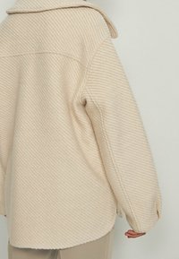 Beige gestructureerde lange mouwen jas met een kraag, gedragen over bijpassende broek, getoond vanuit de achterkant en een beetje van opzij.