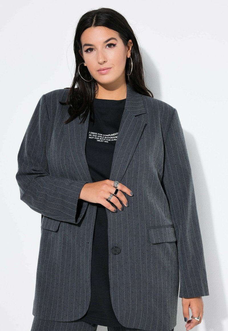 Harmaa raidoitettu bleiseri pitkillä hihoilla, kahdella etutaskulla ja yhdellä nappikiinnityksellä, käytetty mustan graafisen t-paidan päällä. Rohkeaa tekstiiliä.