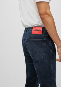 Tmavě modré džínové kalhoty s červenou koženou nášivkou s logem „HUGO“ na zadní straně. Rovný střih s dvěma zadními kapsami.