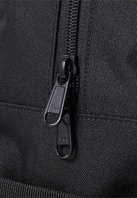 Primer plano de una mochila de tela negra, con un doble cierre YKK, exterior de nylon texturizado y costuras reforzadas a lo largo de las uniones.