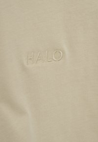 Jasnobeżowa, prążkowana tkanina z haftowanym logo "HALO". Tekstura jest gładka, z delikatnym rozciągliwością i minimalistycznymi elementami wzornictwa.