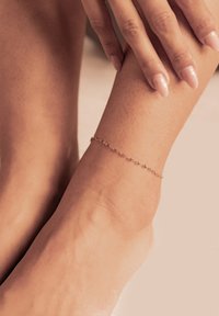Pulsera de tobillo de oro con un diseño de cadena delicada, que presenta pequeños segmentos enlazados. Lleva puesta en un tono de piel suave y claro.
