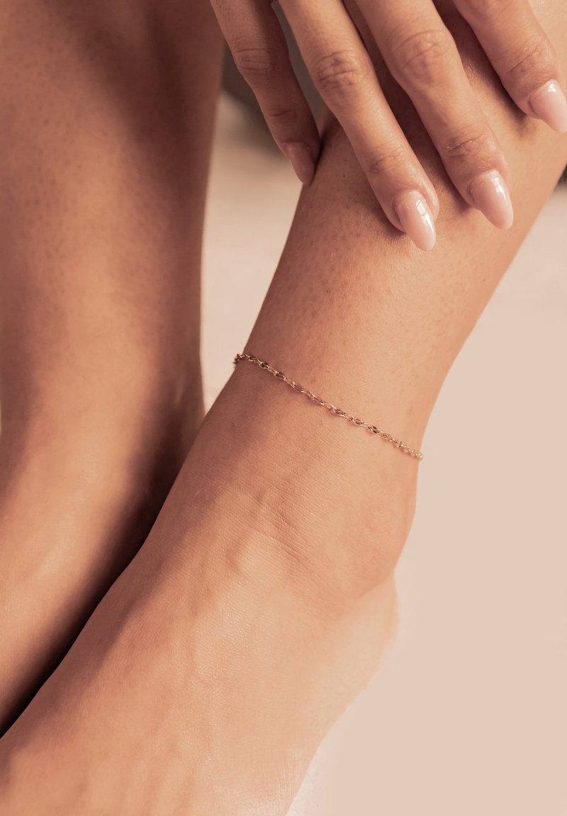 Pulsera de tobillo de oro con un diseño de cadena delicada, que presenta pequeños segmentos enlazados. Lleva puesta en un tono de piel suave y claro.