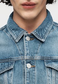 Κοντινό πλάνο ενός ατόμου που φοράει ανοιχτό μπλε τζιν μπουφάν με ασημένια κουμπιά και δύο τσέπες στο στήθος, με το γιακά κλειστό.