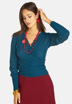Frau trägt einen türkisfarbenen Wickelpullover, roten Rock, rote Blumenohrringe, rote Perlenkette mit Blumendetail und goldene Armreifen, berührt mit einer Hand ihr Haar.