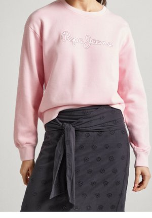 Kobieta w jasno-różowej bluzie z napisem "Pepe Jeans" i ciemnoszarej spódnicy z kwiatowymi wycięciami, związanej w talii.