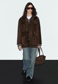Persona che cammina indossando una giacca oversize marrone scuro, jeans blu, scarpe nere, grandi occhiali da sole scuri e portando una borsa marrone abbinata.