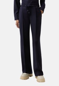 Persoon in navyblauwe rechte broek met elastische tailleband en beige schoenen met dikke zool, hand in zak, tegen een effen achtergrond.