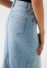 Heledsinine denim seelik, mis on alati keha lähedal, tagaküljel tasku, millel on kolmnurkne punane 'GUESS' logo ja nähtavad õmblusdetailid.