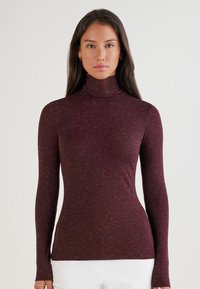 Braunrote hochgeschlossene Langarmbluse mit strukturiertem Finish, in slim fit und feinem gesprenkeltem Muster, kombiniert mit weißen Hosen.