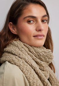 Beige neulottu huivi, jossa on teksturoitu kuvio, ripustettuna löysästi kaulan ympärille, valmistettu pehmeästä materiaalista, jota täydentää kevyt ulkokerros.