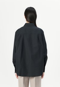Fekete, nagyméretű ing klasszikus gallérral, hosszú ujjal és gombos mandzsettával. Sima textúra, minimalista design, diszkrét márkajelzéssel a hátsó részen.