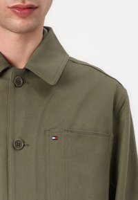 Oliven grønn stoffskjortejakke med kragehals, som har to knapper og en brystlomme med en liten logo-detalj.