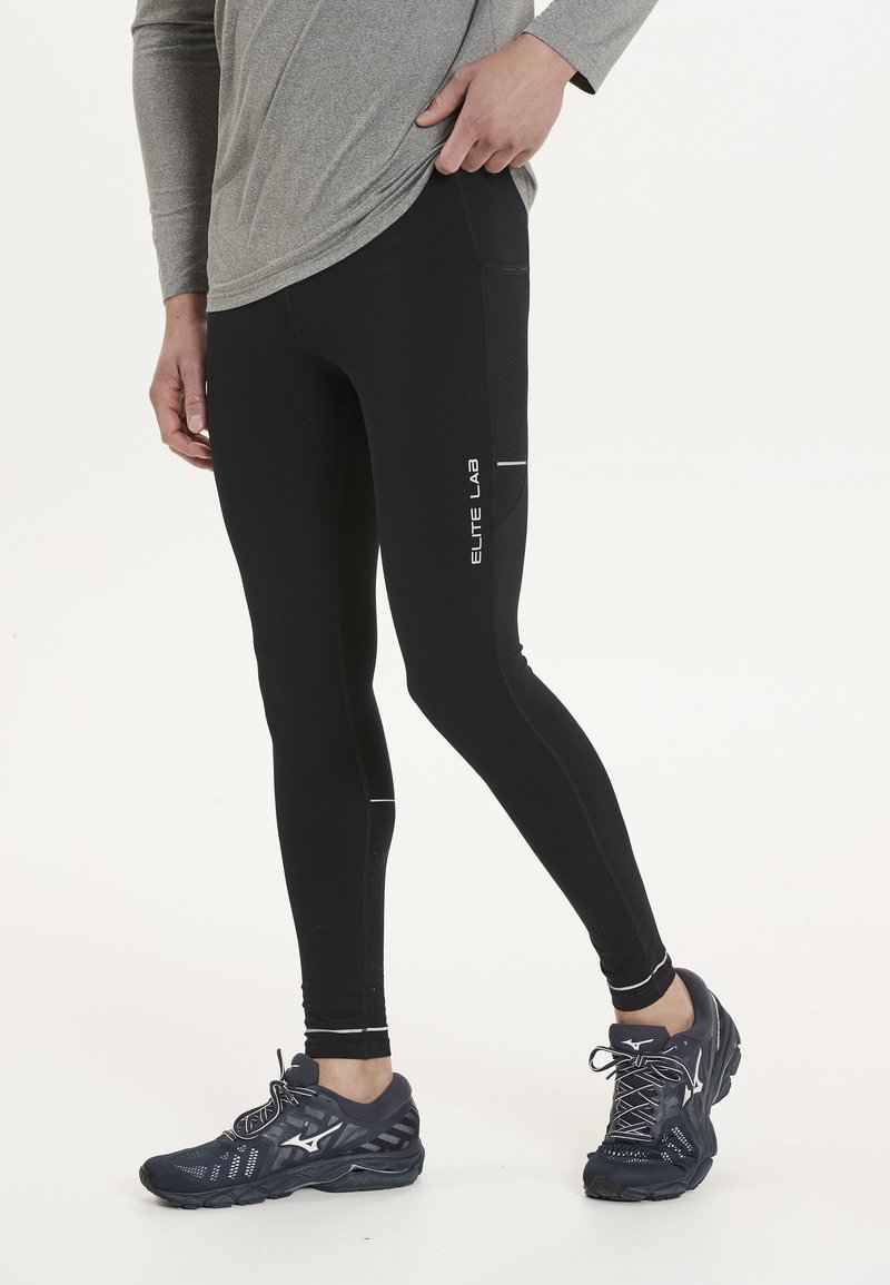Zwarte sportlegging met een aansluitend ontwerp, voorzien van mesh accenten en reflecterende details. Gecombineerd met marineblauwe hardloopschoenen.