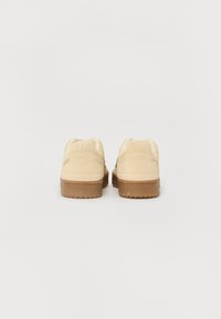 Beige suede sneakers med en brun gummisåle. Har polstrede ankelkraver og subtile grønne detaljer på sidene. Minimalistisk design.
